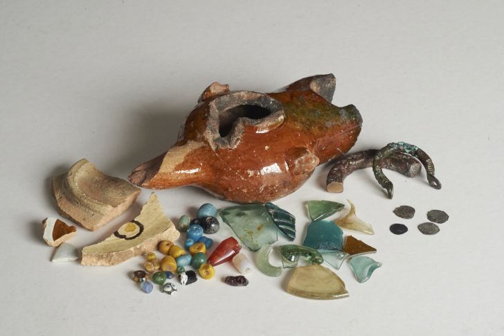A selection of excavated finds from Essouk-Tadmekka, including fragments of glazed ceramics, stone beads, a cowrie shell, a fragment of silk textile, a carved stone torso, and vessel glass fragments.