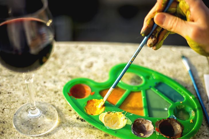 A painter dips a brush into a palette that sits next to a glass of red wine