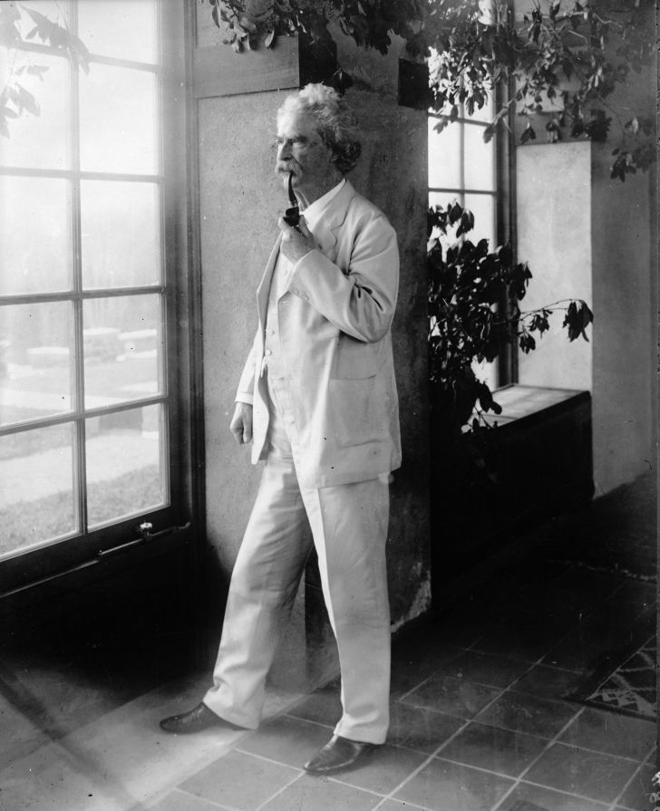 Mark Twain standing near a window while wearing a white suit and smoking a pipe