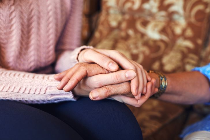 A person in a pink sweater, sitting on a couch, holding the hands of an older person