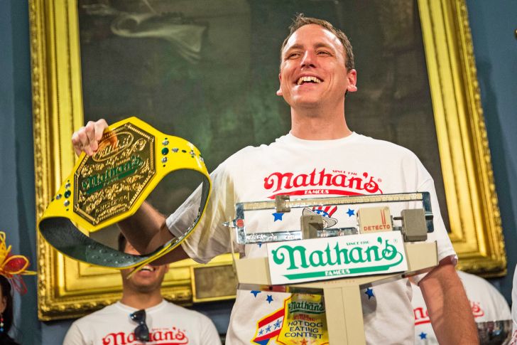 Image of Joey Chestnut standing on a scale and holding the prize-winning mustard yellow bet for winning the hot dog eating competition