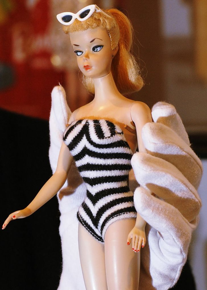 A gloved hand holding a vintage Barbie doll wearing a black and white knit bathing suit.
