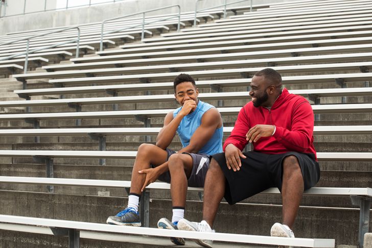 Two black men in sporty clothing sitting on bleachers and laughing