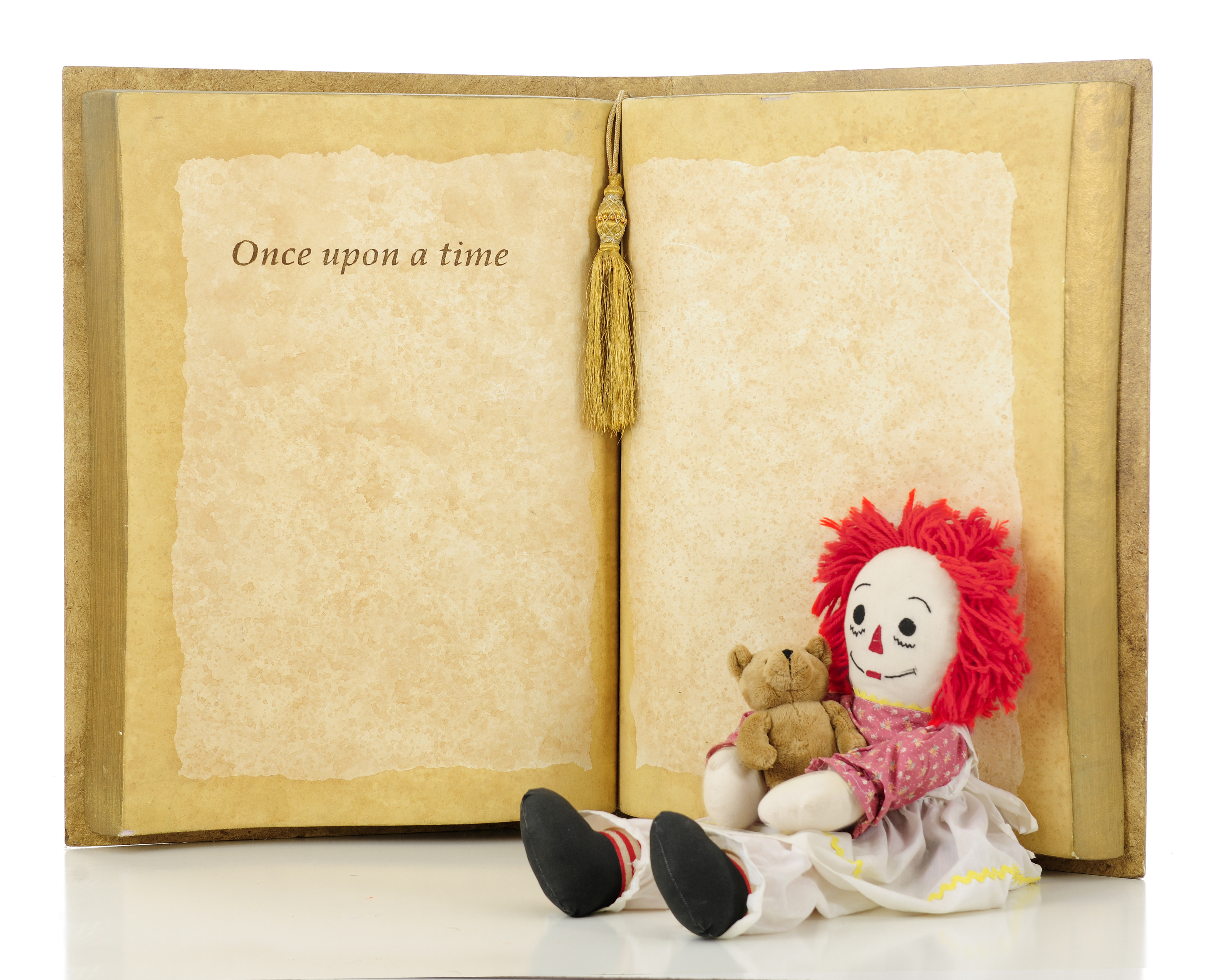A rag doll with red yarn hair and holding a tiny teddy bear is sitting in front of an open book that reads "Once upon a time..."